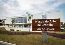 Fotografia da fachada contemporânea do Museu de Arte de Brasília com placa de sinalização bilíngue (português e inglês), vegetação paisagística e céu parcialmente nublado ao fundo.