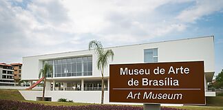 Fotografia da fachada contemporânea do Museu de Arte de Brasília com placa de sinalização bilíngue (português e inglês), vegetação paisagística e céu parcialmente nublado ao fundo.